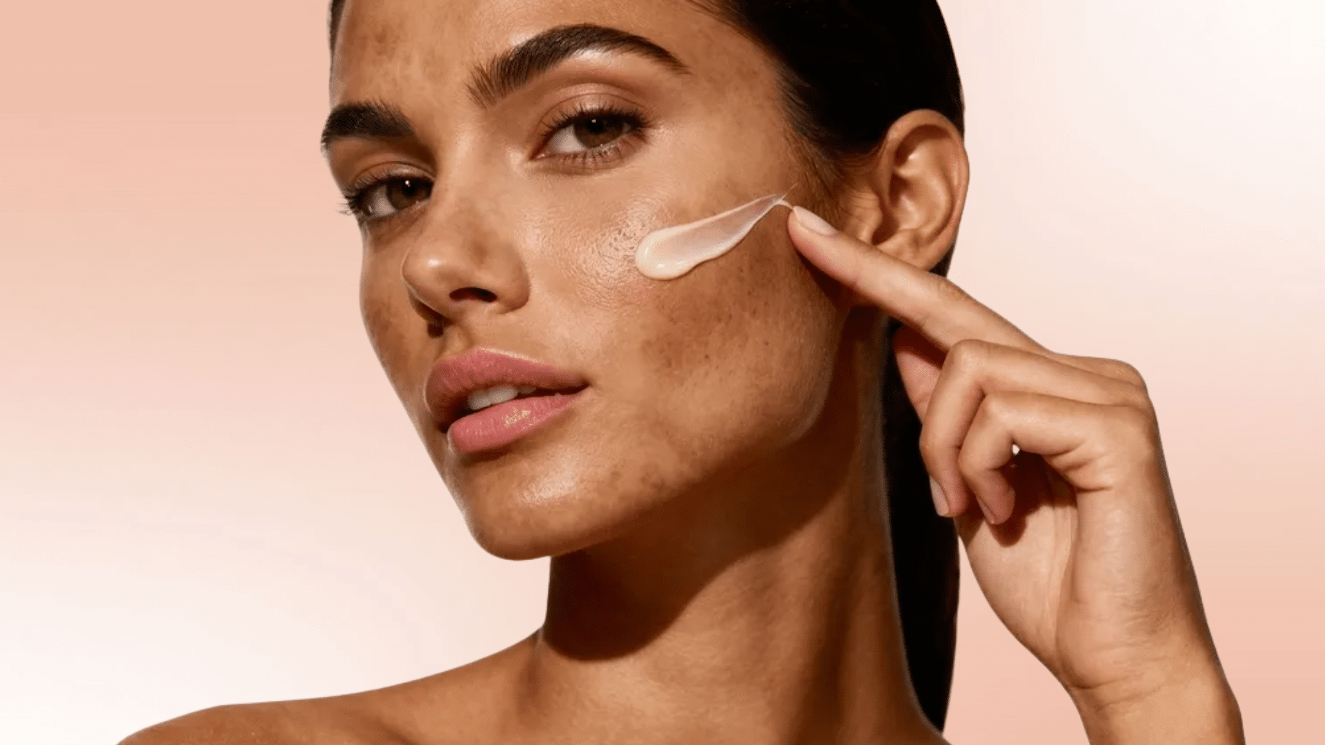 Woman applying cream on cheek with fingertip against a soft pink background, close-up skincare application focused on skin texture and tone.