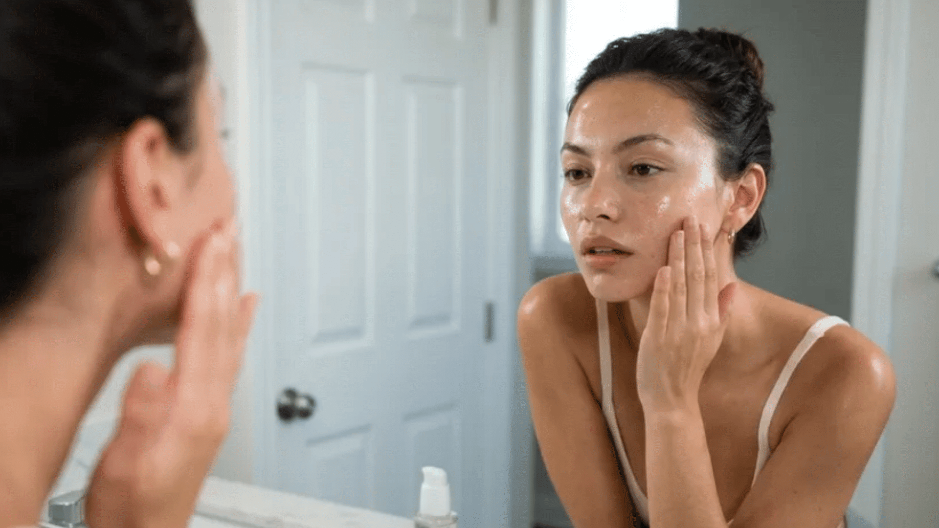 Woman applying skincare while looking in bathroom mirror, touching cheek, serum bottle on bathroom counter