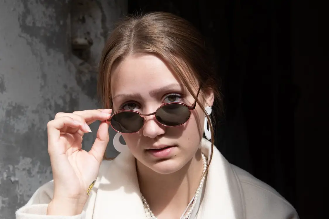 Woman in a light coat adjusting sunglasses indoors with a textured wall background