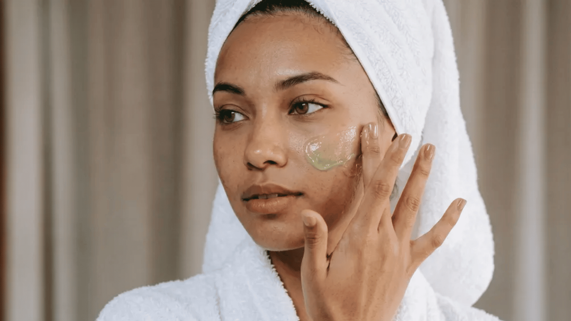 Woman in towel applying green skincare gel to cheek while looking slightly sideways during skincare routine