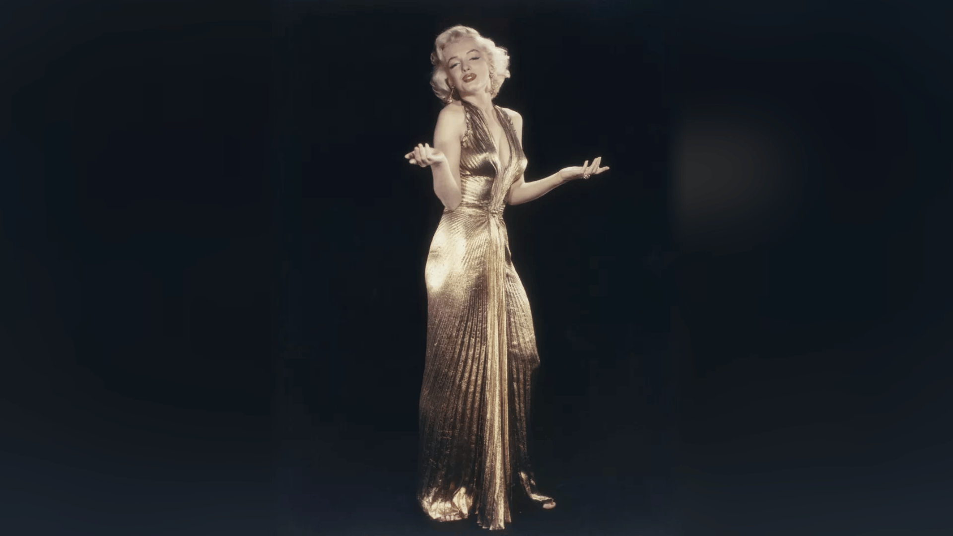 Marilyn Monroe posing in a pleated gold halter gown against a dark studio backdrop