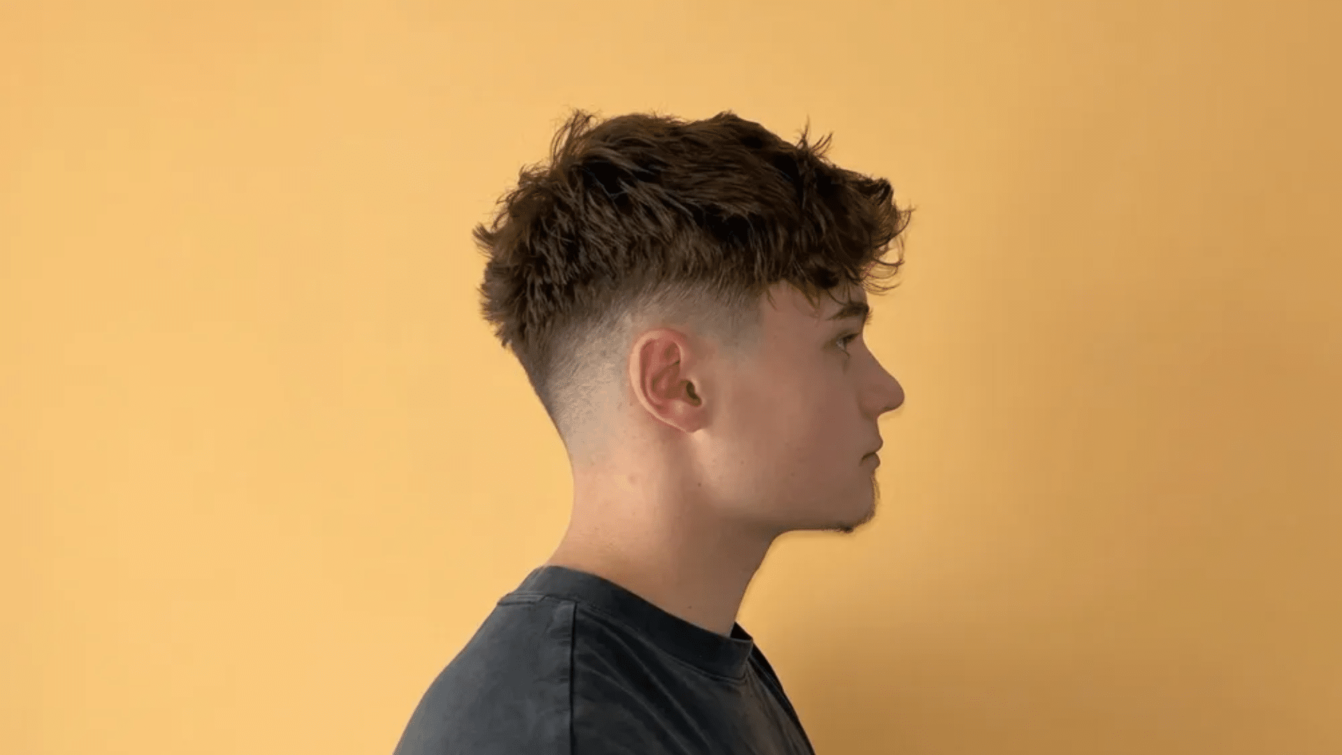 Young man in side profile with short mullet haircut, featuring faded sides, textured top, and slightly longer back against a warm background.