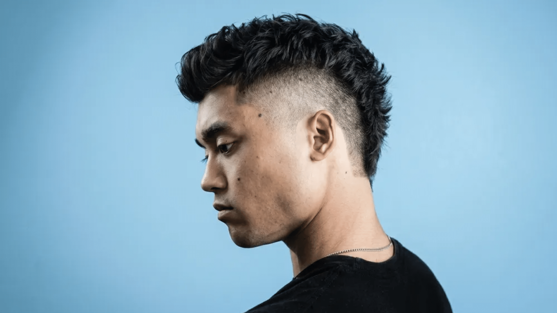 Young man in side profile with short mullet haircut, featuring high faded sides, textured top, and longer back section in a clean blue studio setting.