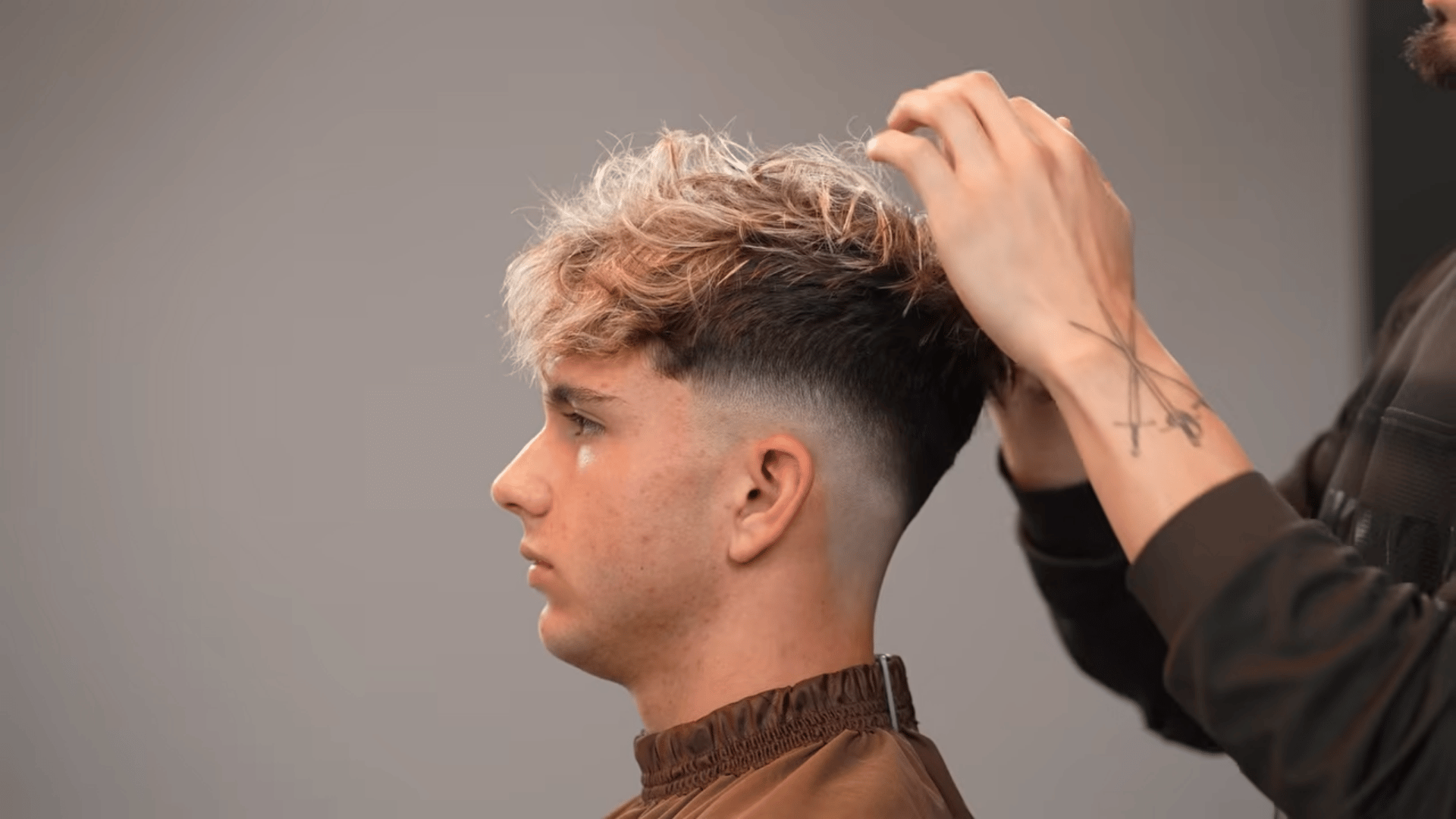 A barber using his hands to style the textured, blonde-highlighted top of a client's finished skin fade haircut.