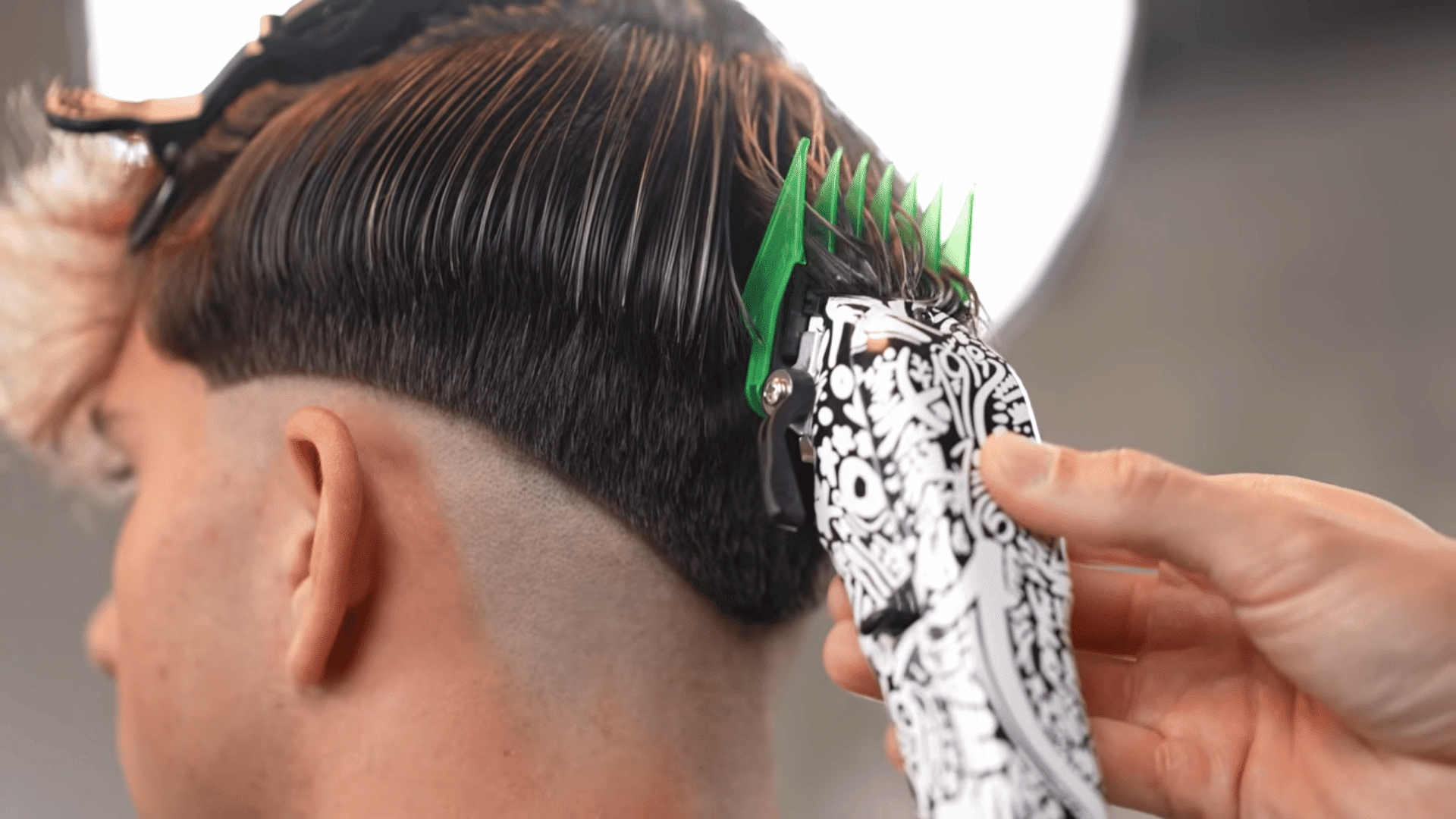 A barber using patterned clippers with a green guard to blend and fade a client's dark hair.