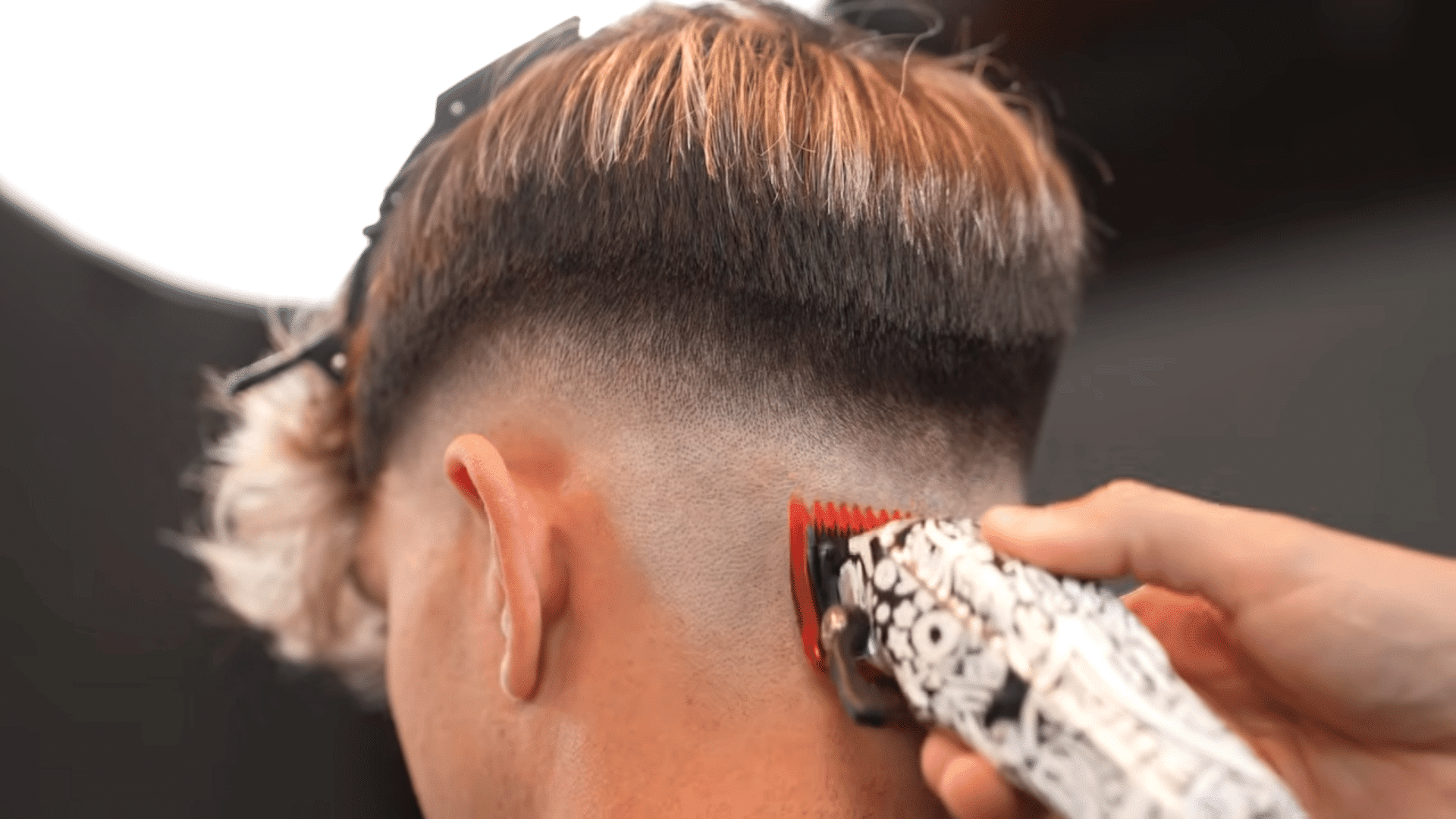 A barber using patterned clippers with a red guard to shave and fade the lower neckline of a client's haircut.
