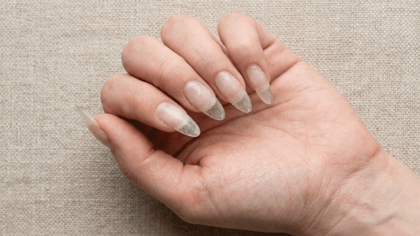 Palm-up view of fingers with long, clear pointed acrylic nails on woven fabric.
