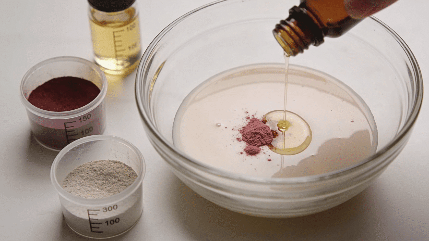 Pigment, mica, and flavor oil being added in small amounts to a cosmetic mixture in a bowl