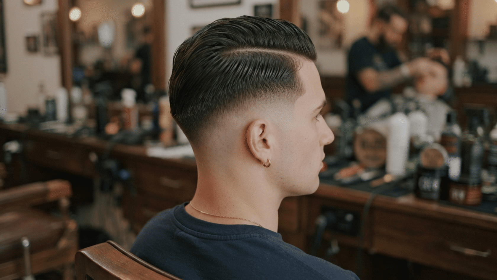 Back side profile of man with slicked back haircut, low fade, hoop earring, gold necklace, black shirt and blurred background in a barber's shop