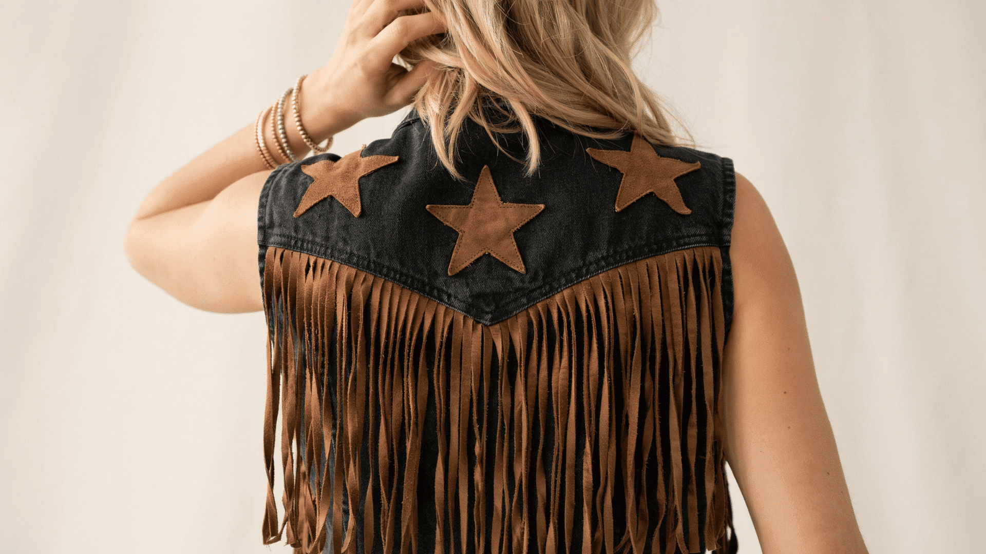 Back view of a black denim boho festival vest featuring brown suede stars, long hanging fringe, and gold bracelets