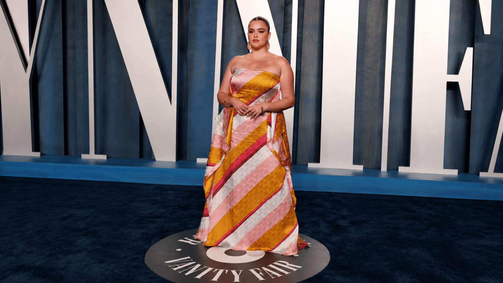 Barbie Ferreira wearing a colorful strapless gown on a red carpet with a dark geometric backdrop.