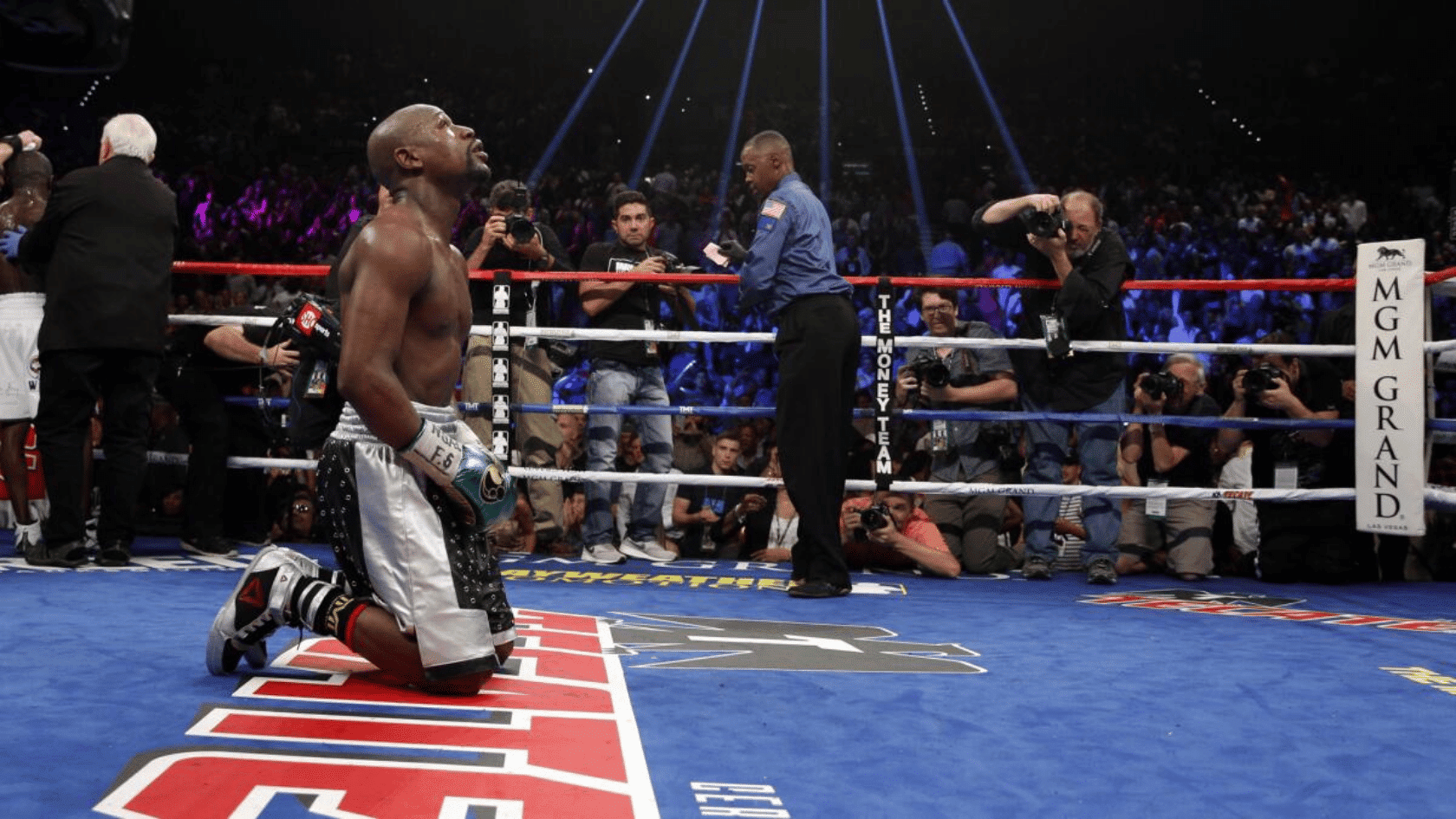 Boxer kneeling in boxing ring after bout, surrounded by photographers and officials, crowd in arena, ropes visible