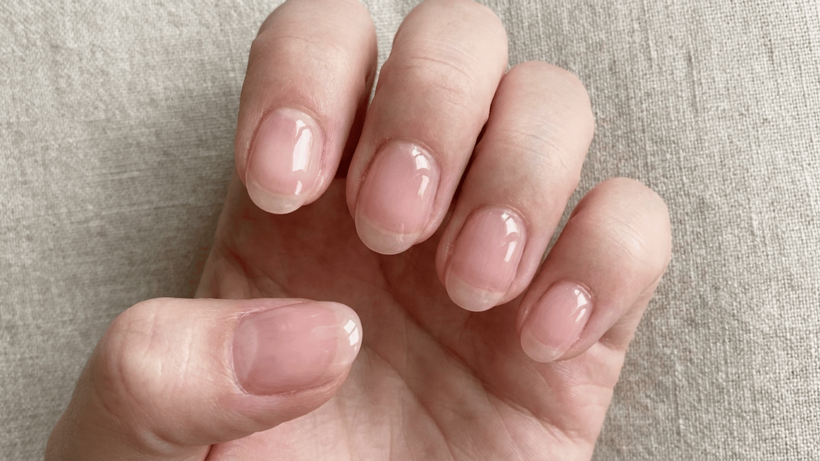 Palm-up view of fingers with glossy, sheer pink builder gel nails on a woven fabric surface.