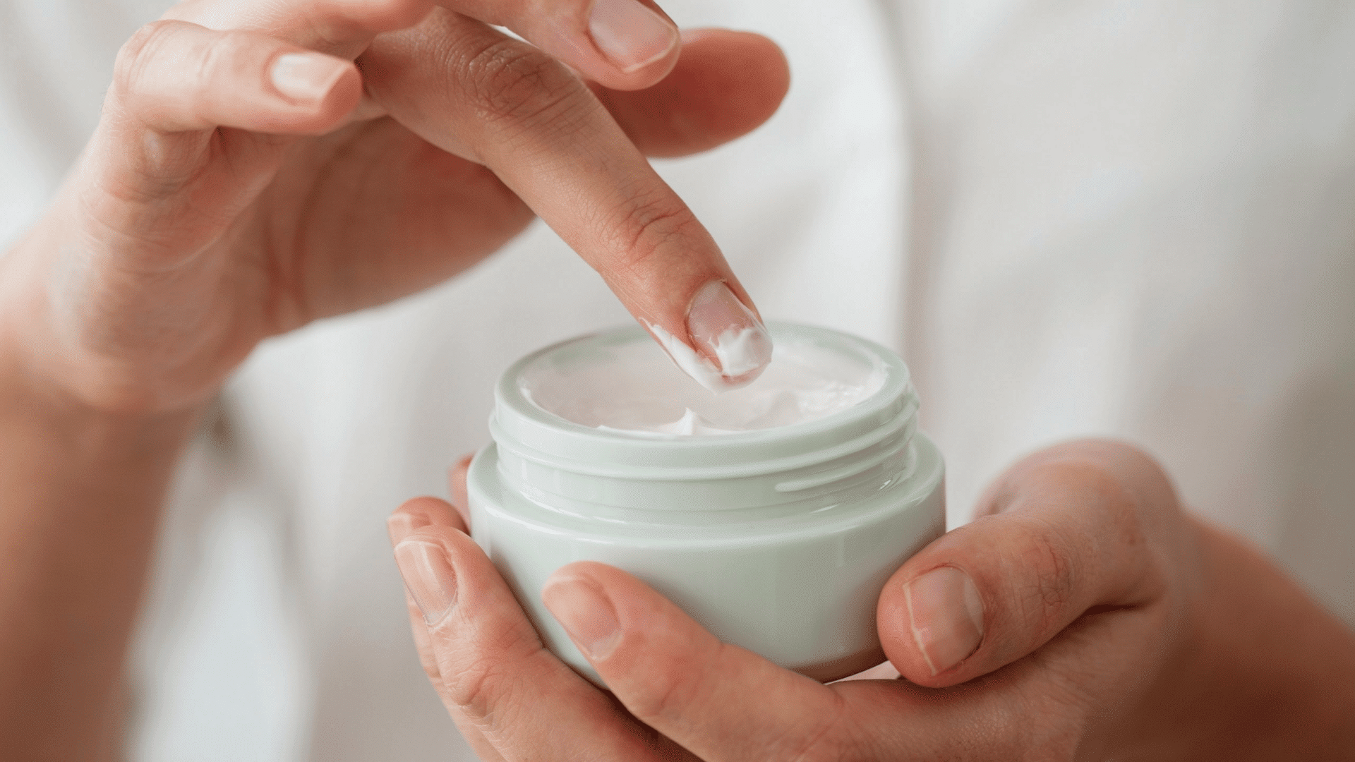 Close-up of hands holding an open jar and dipping a finger into white moisturizing cream