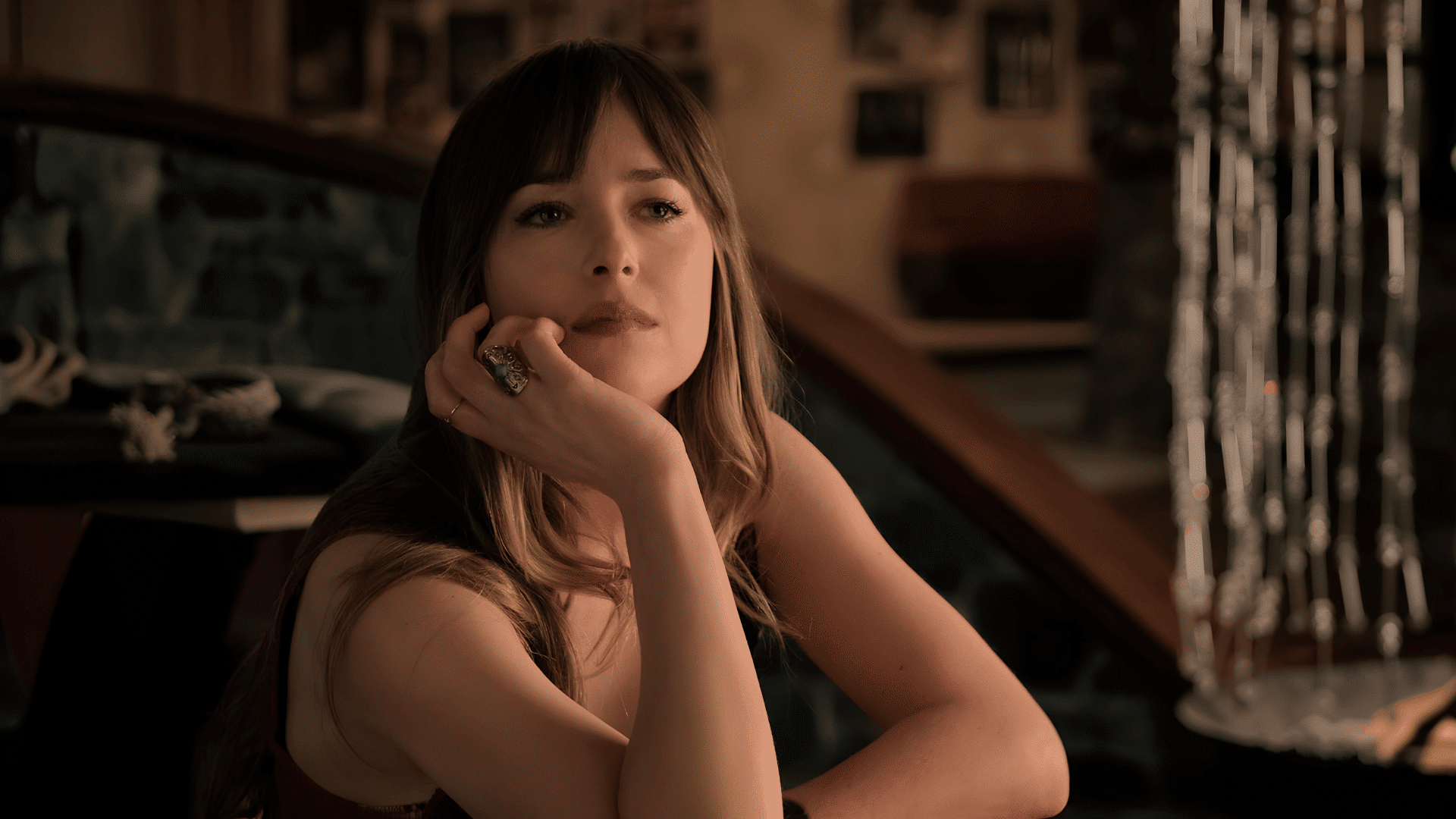 Dakota Johnson with long piecey bangs framing her heart-shaped face as she rests her chin on her hand.