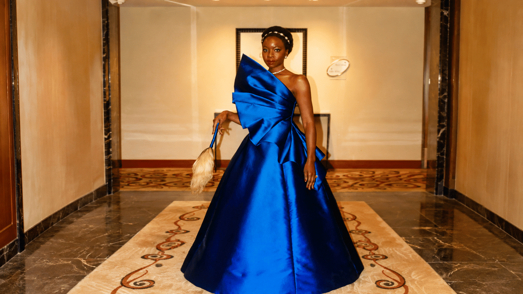 Danai Gurira wearing a blue one-shoulder ball gown in a hotel hallway setting.