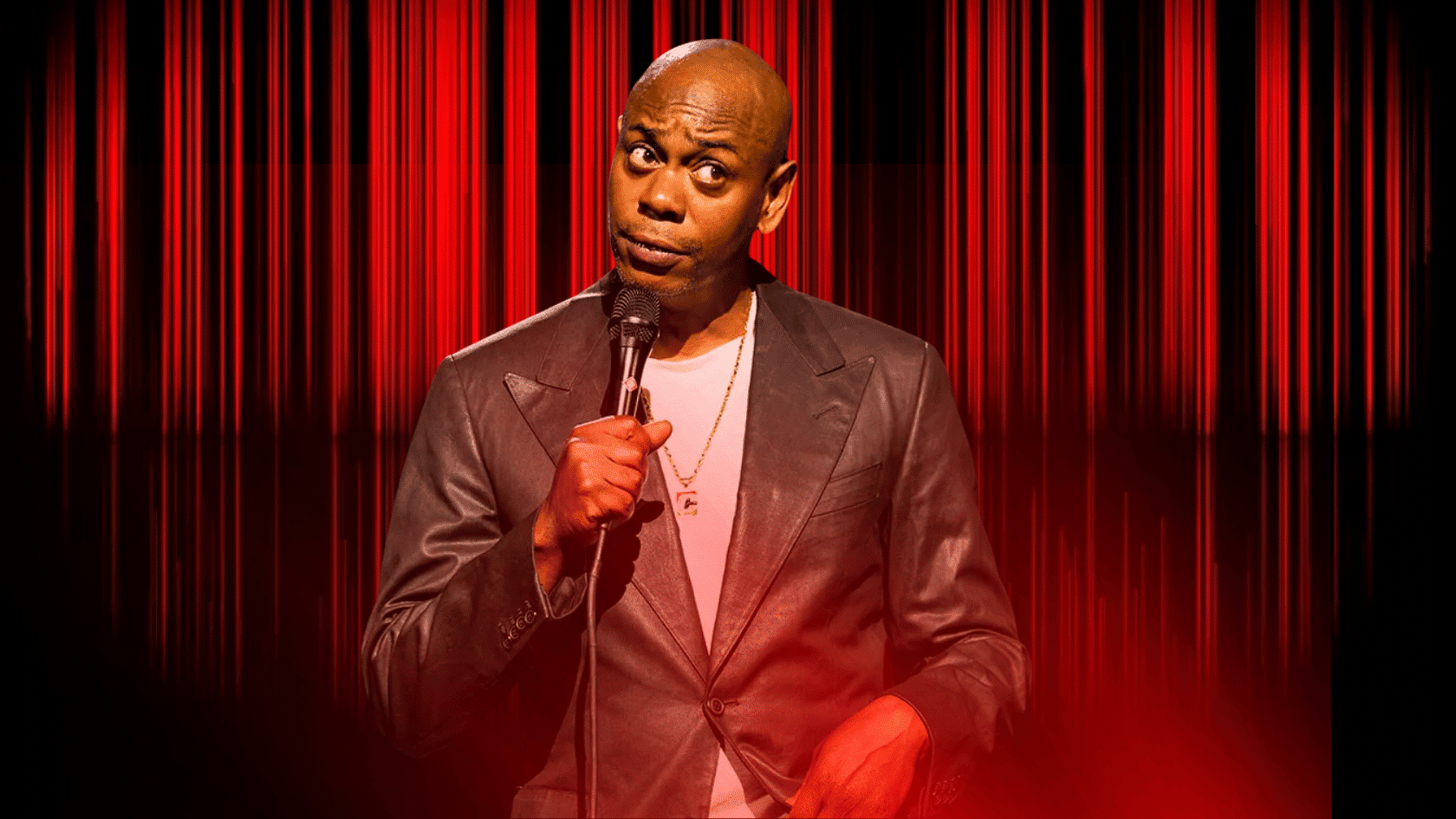 Dave Chappelle performing on stage with a microphone against a red curtain background.