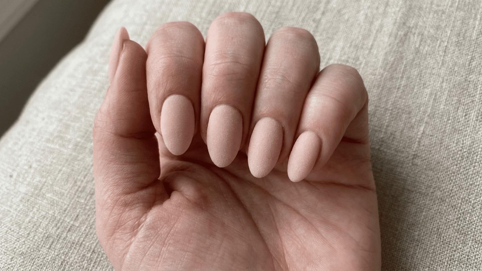 Close-up view of oval-shaped nails with matte nude gel polish on a hand.