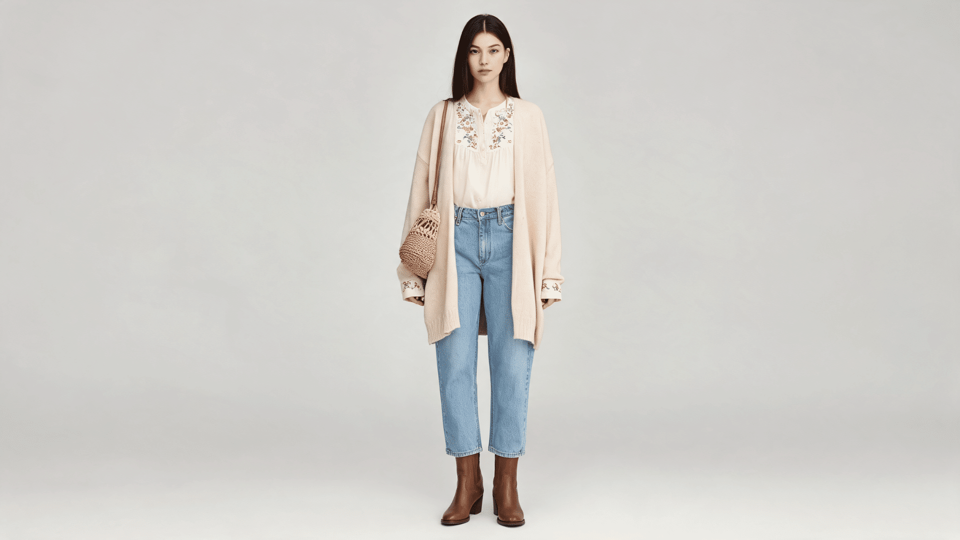 Female model wearing relaxed jeans, embroidered blouse, ankle boots, cardigan, and a woven crossbody bag