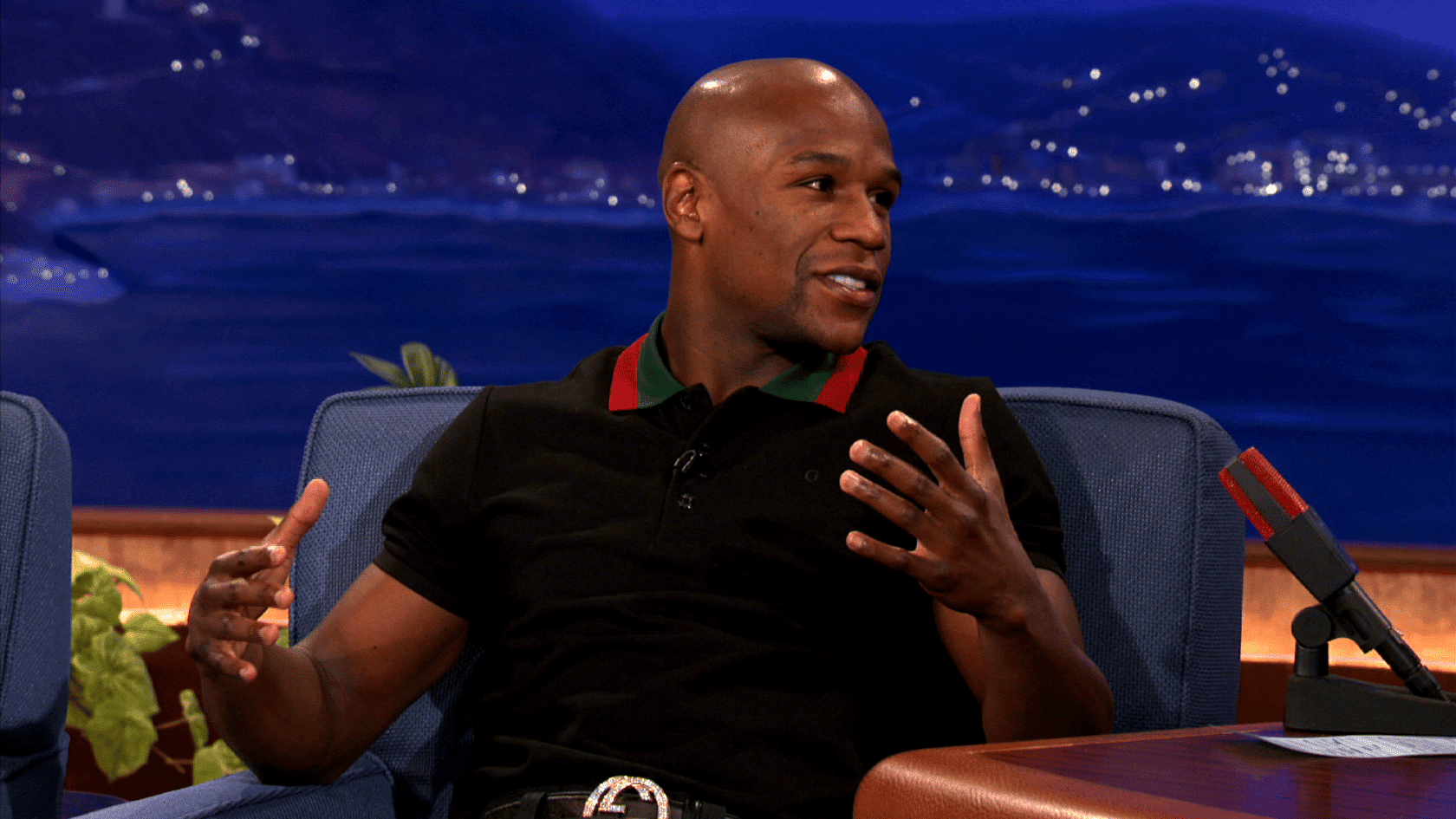 Floyd Mayweather seated on talk show couch speaking wearing black polo shirt, blue studio background