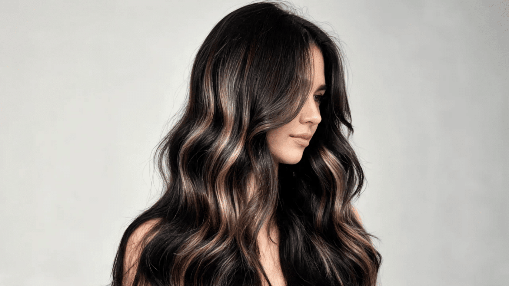 Woman with long black hair featuring brown highlights, styled in waves with a side profile against a plain background.
