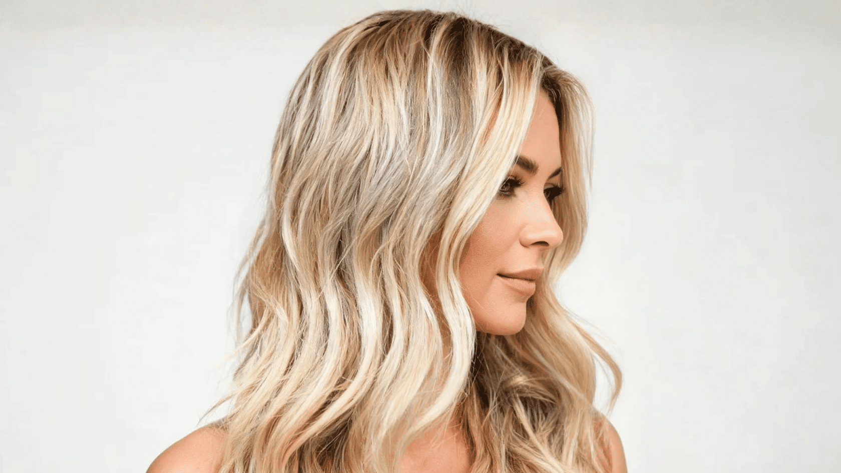 Woman with long blonde highlighted hair styled in loose waves, shown in side profile against a plain background.