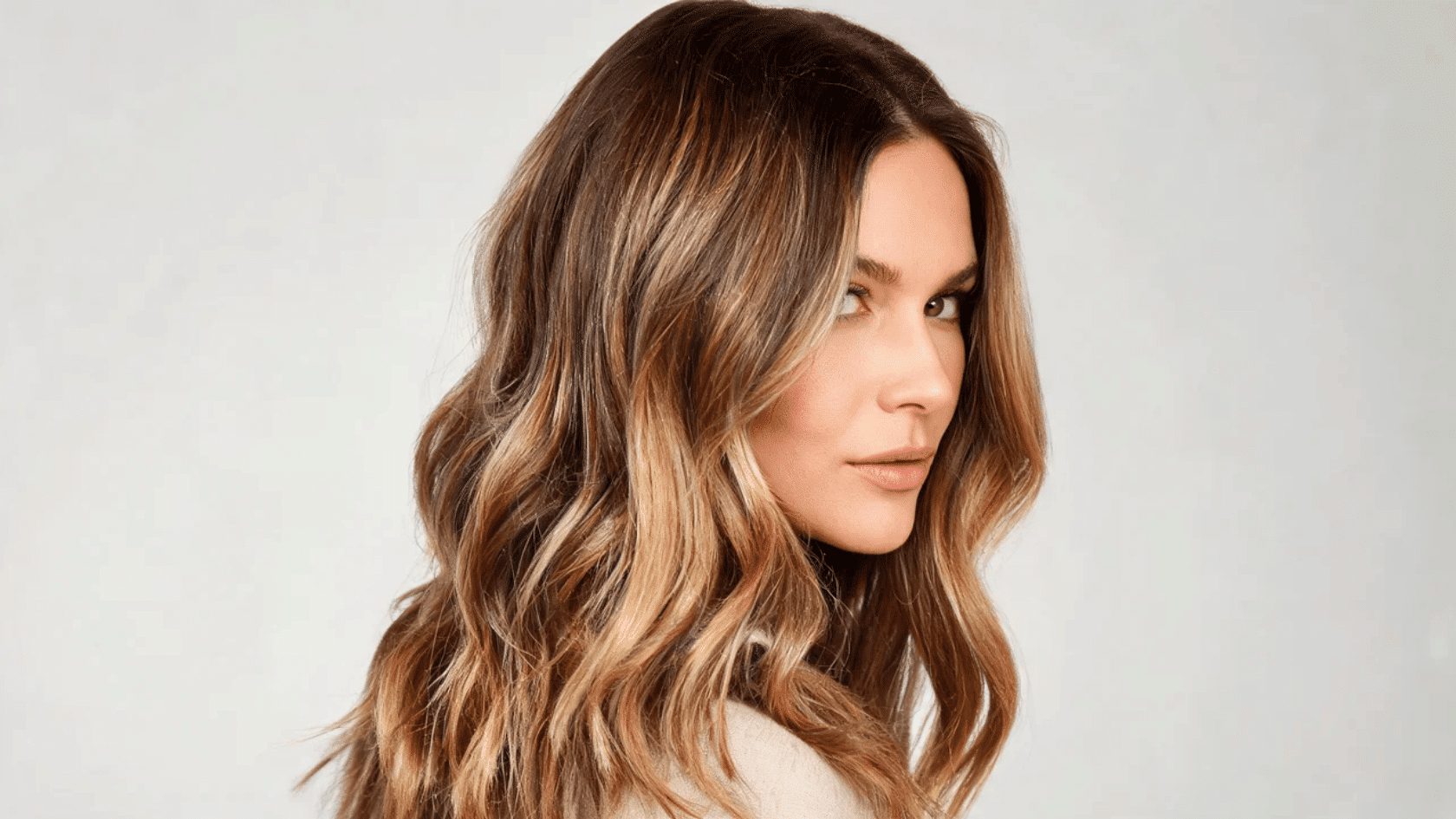 Woman with long brown highlighted hair styled in waves, facing sideways against a plain background.