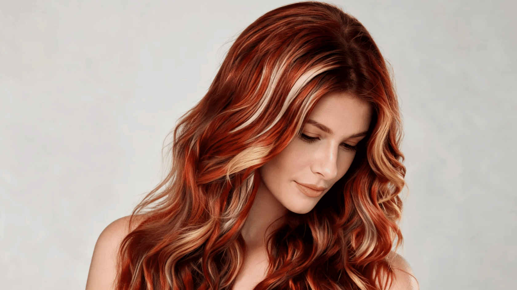 Woman with long red highlighted hair styled in waves, looking downward against a plain background.