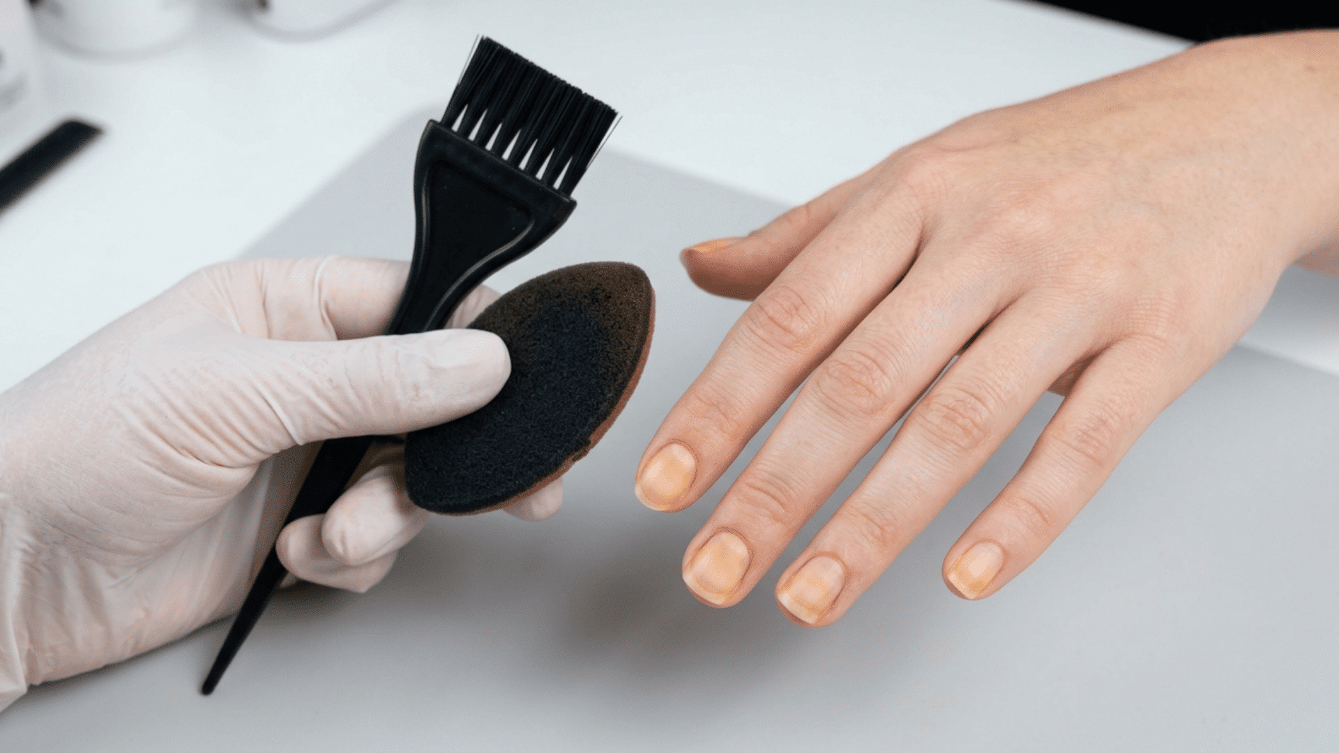 Gloved hands holding hair dye and self-tanner applicators beside lightly stained natural fingernails.