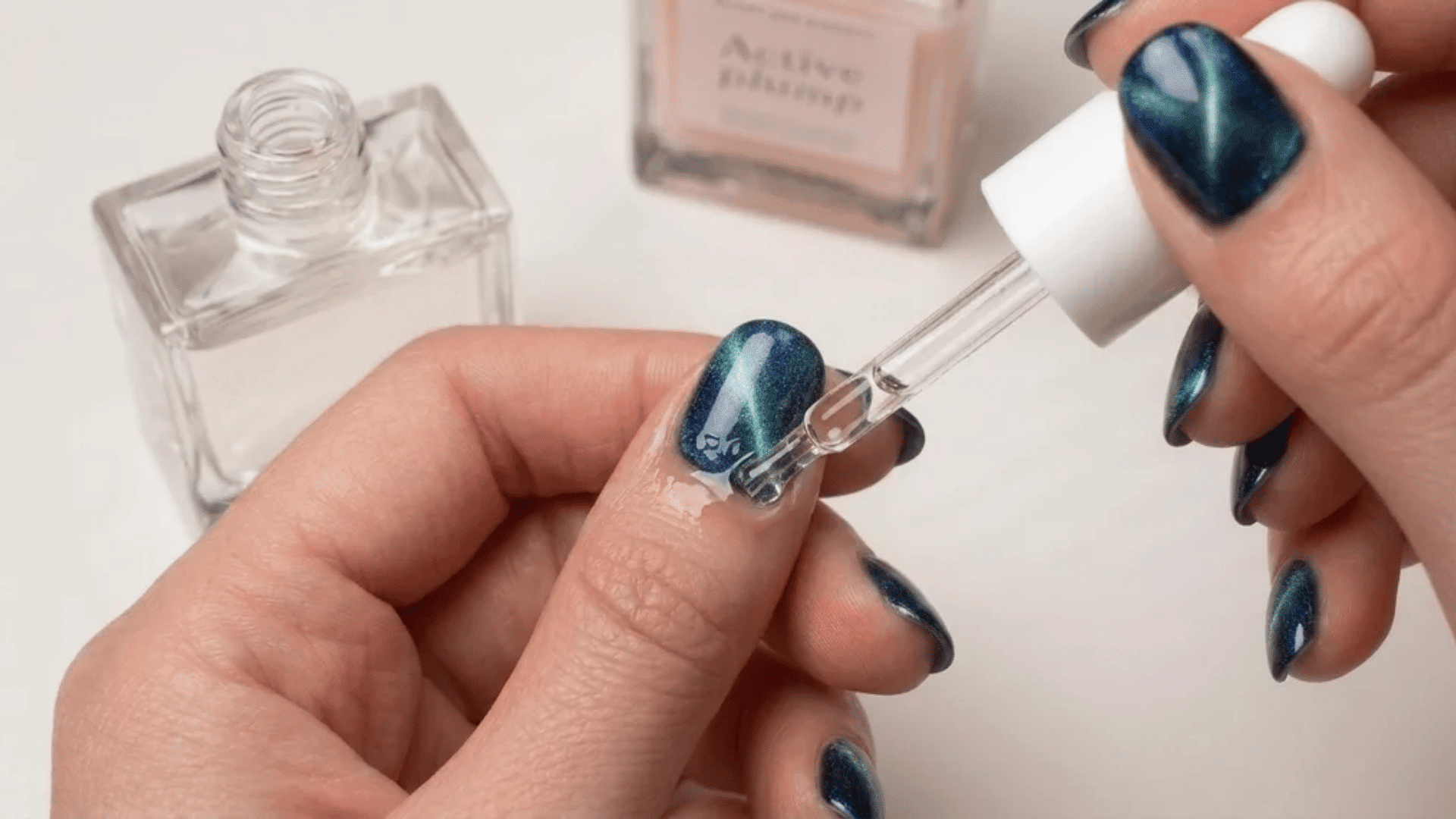 hand applying cuticle oil to blue-green nail using dropper with nail polish bottles in background