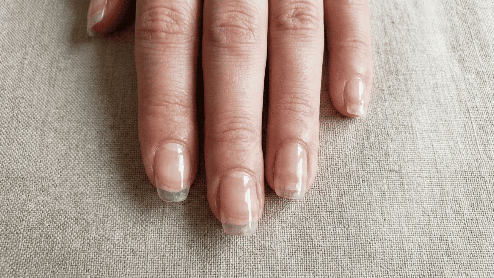 Close-up of fingers with sheer nude hard gel nails, resting on a woven fabric surface.