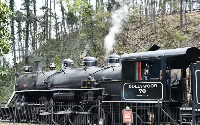 Black steam locomotive with "Dollywood" logo emitting steam in wooded outdoor setting
