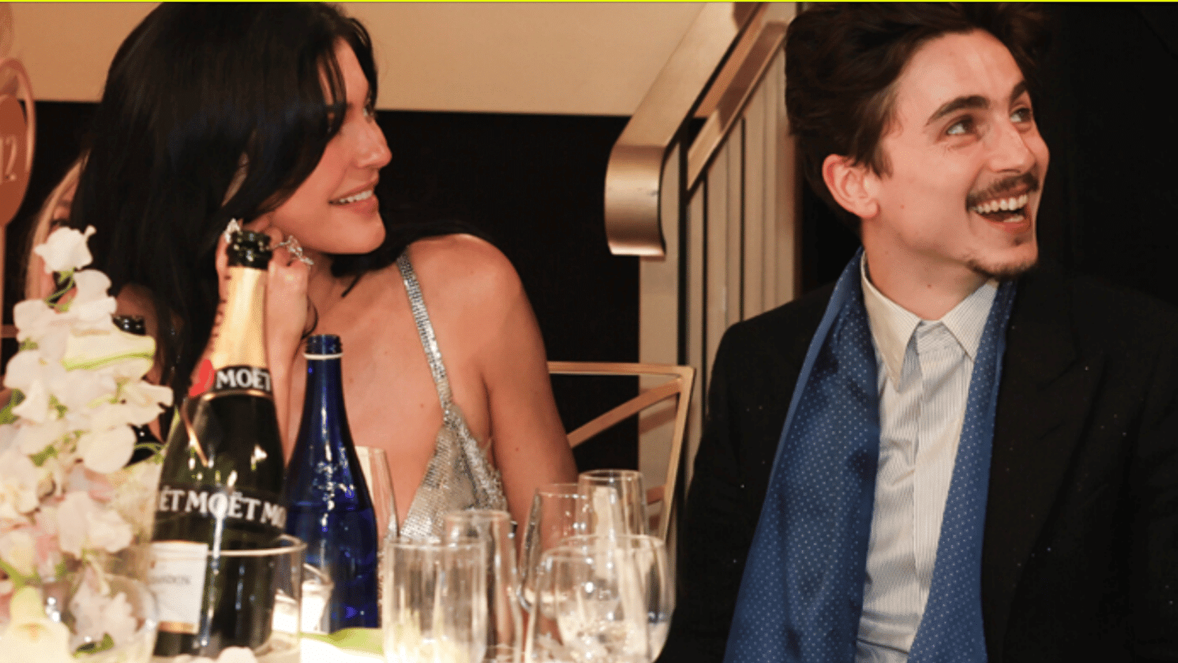 Kylie Jenner and Timothée Chalamet smiling at a dinner table with champagne glasses