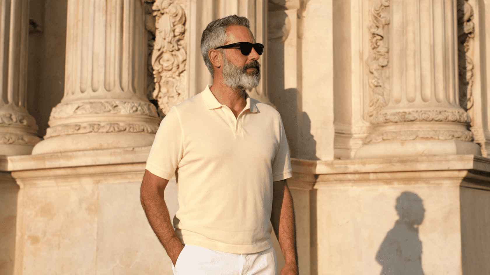 Man wearing a white polo shirt and matching trousers standing outdoors beside large stone columns.