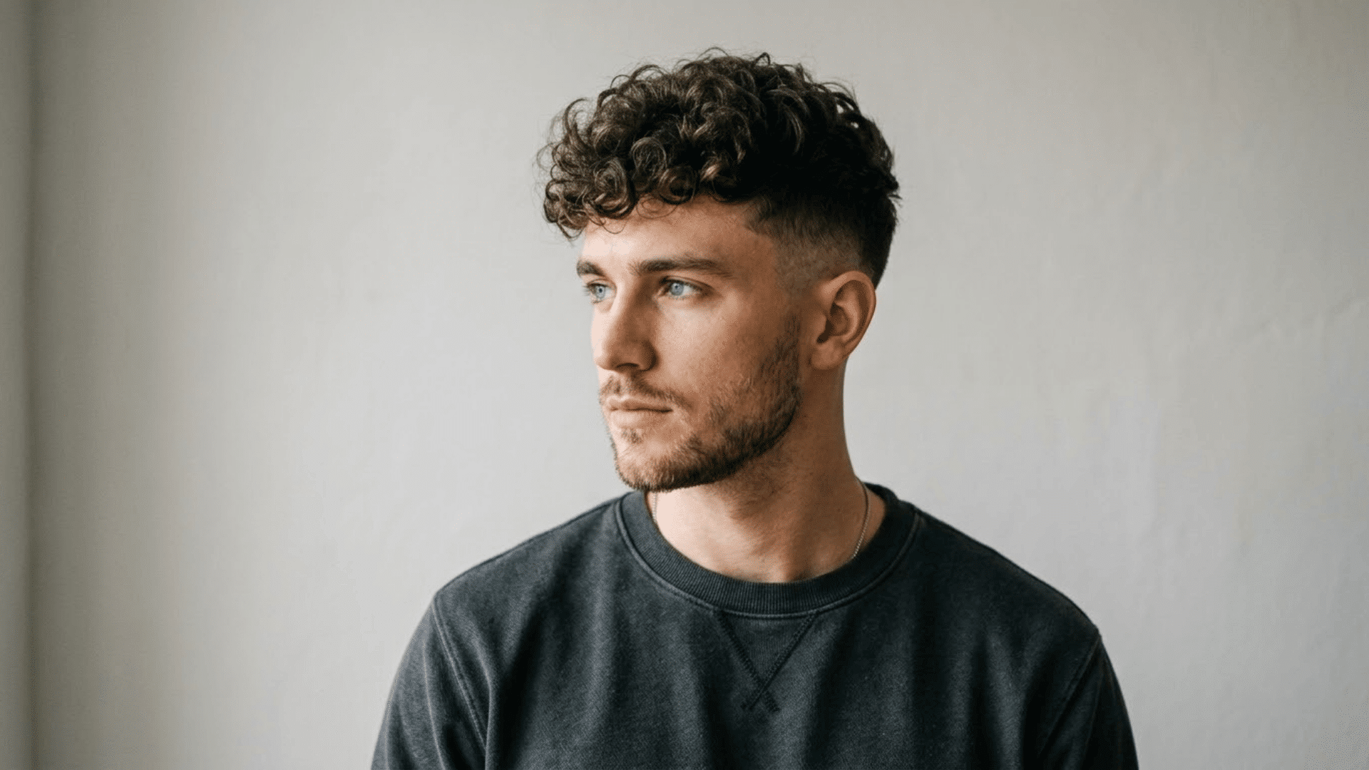Man with a curly crop fade haircut wearing a dark sweatshirt against a plain light background.