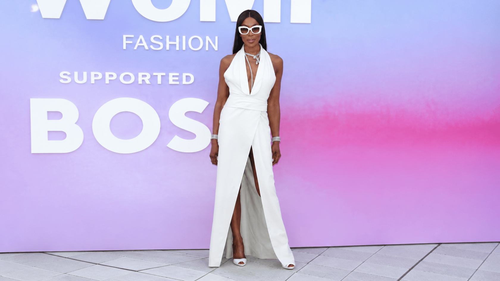 Naomi Campbell wearing a white halter gown and sunglasses at a fashion event with a pastel backdrop.