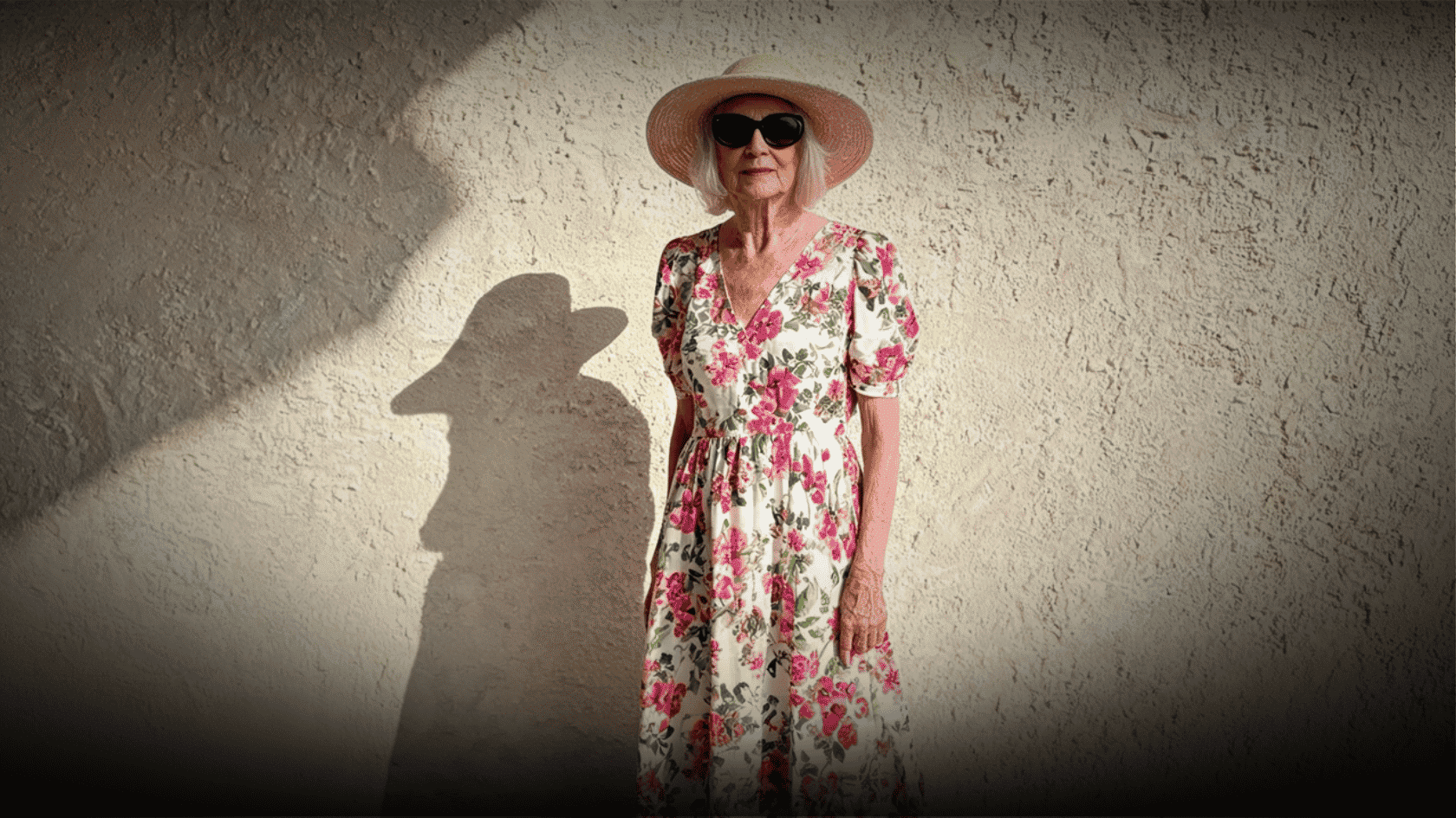Older woman wearing a floral boho dress, sun hat, and sunglasses outdoors.