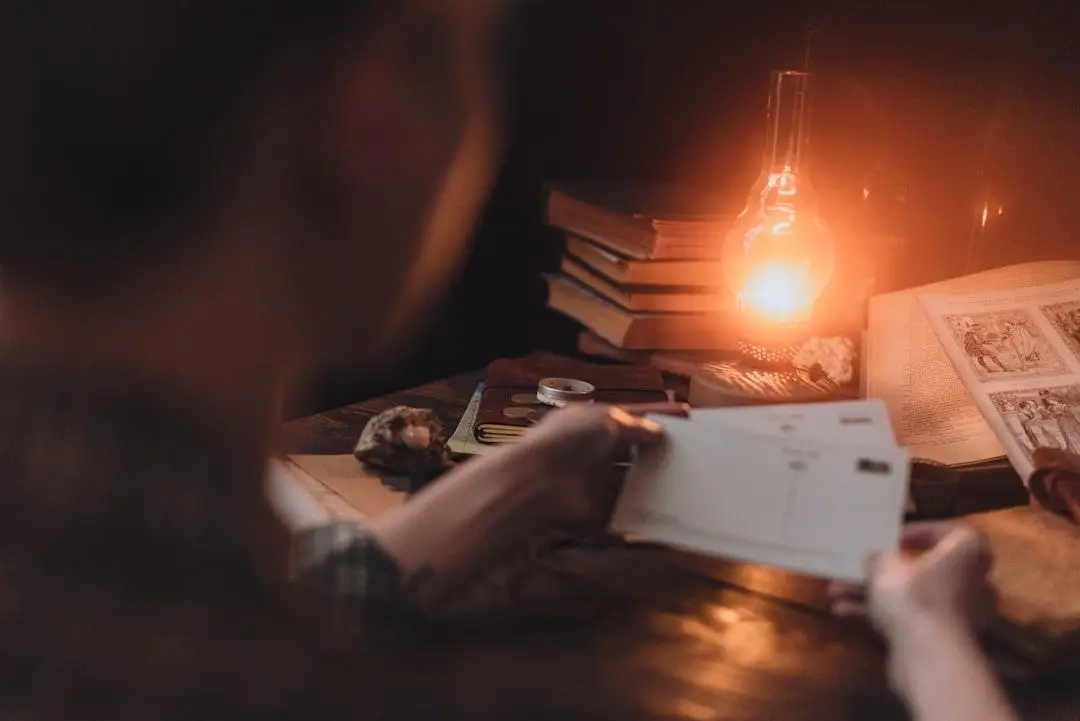 Person reading postcards by dimly lit oil lamp on wooden table with books and papers