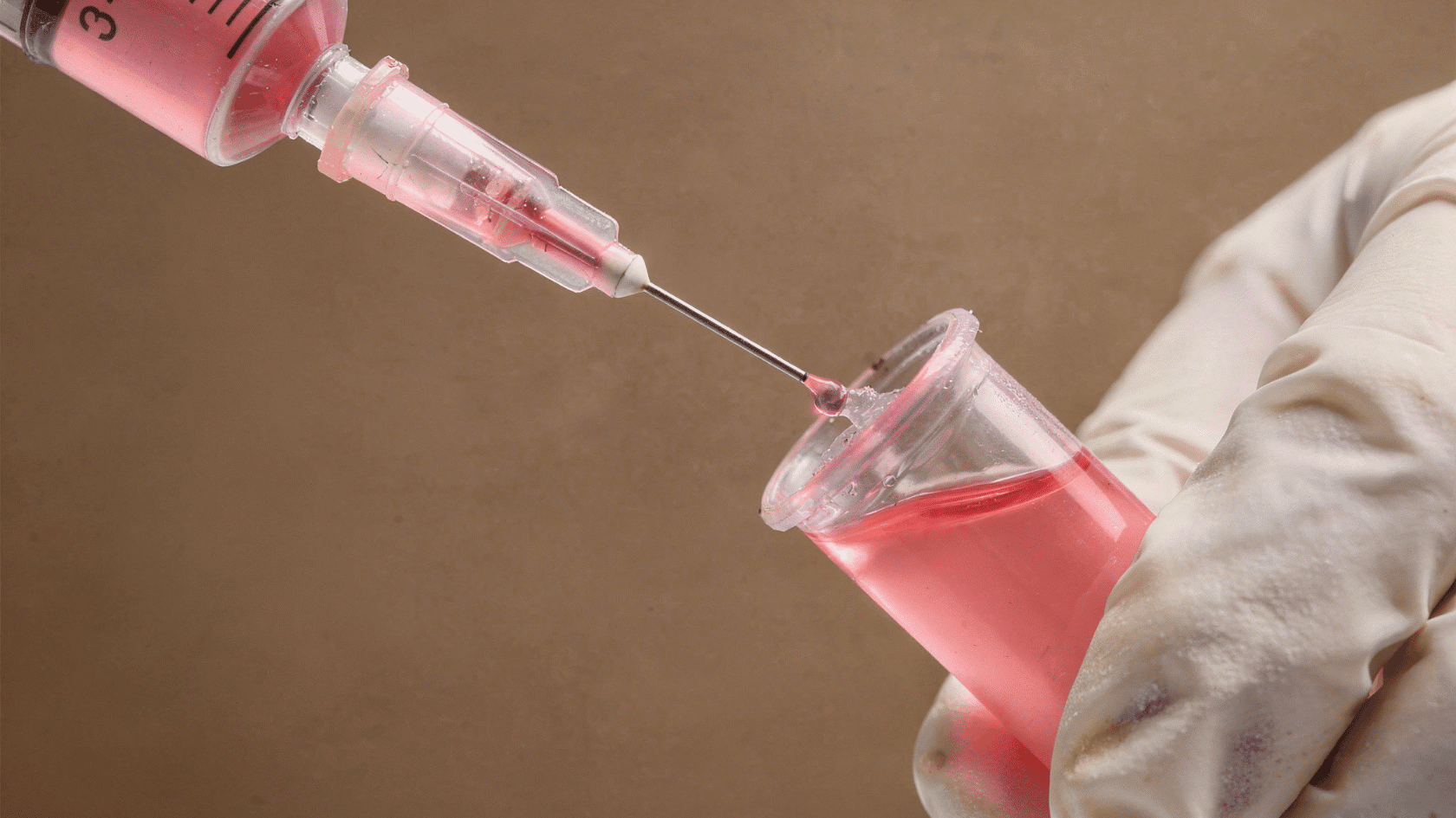 Gloved hand injecting pink liquid into a small lab beaker using a syringe pipette