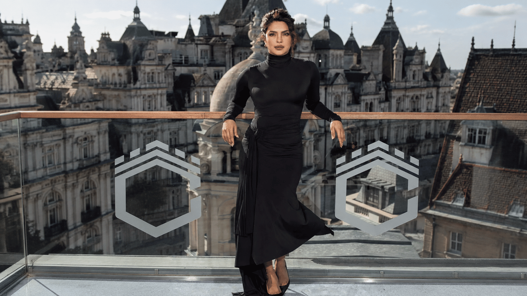 Priyanka Chopra wearing a black long-sleeve gown on a rooftop terrace with a cityscape background.