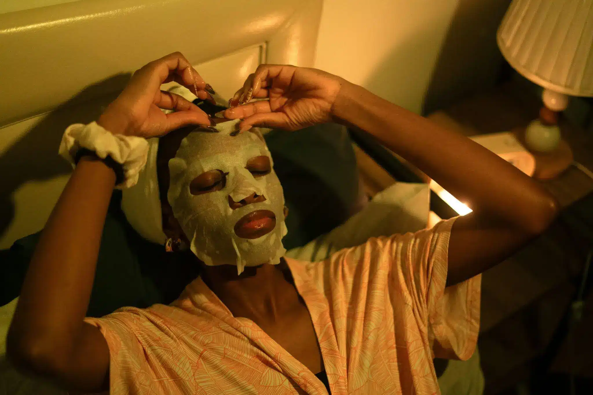 Person relaxing with a face mask in dimly lit bedroom next to a lamp