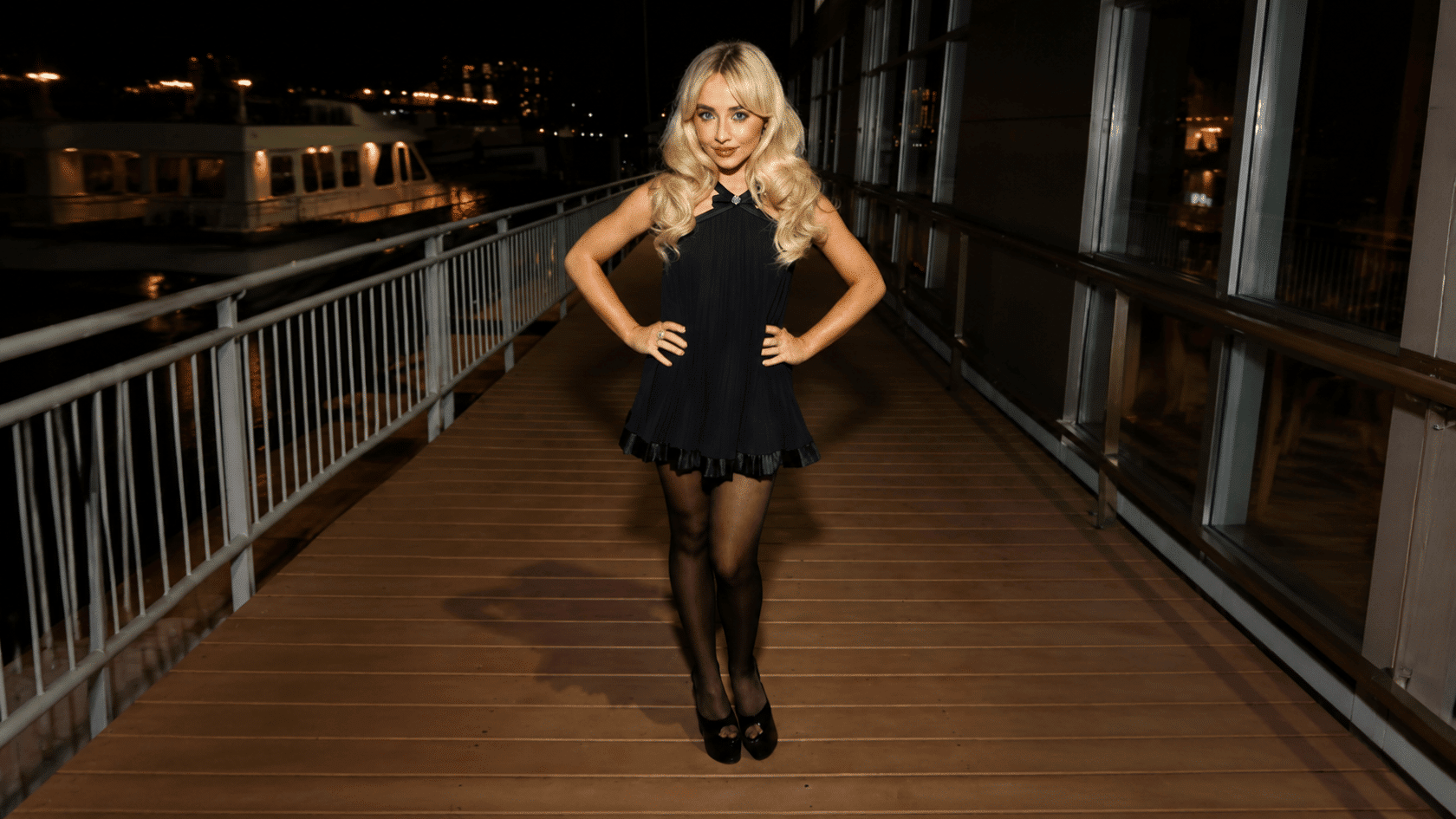 Sabrina Carpenter wearing a black dress and tights on a wooden boardwalk at night.