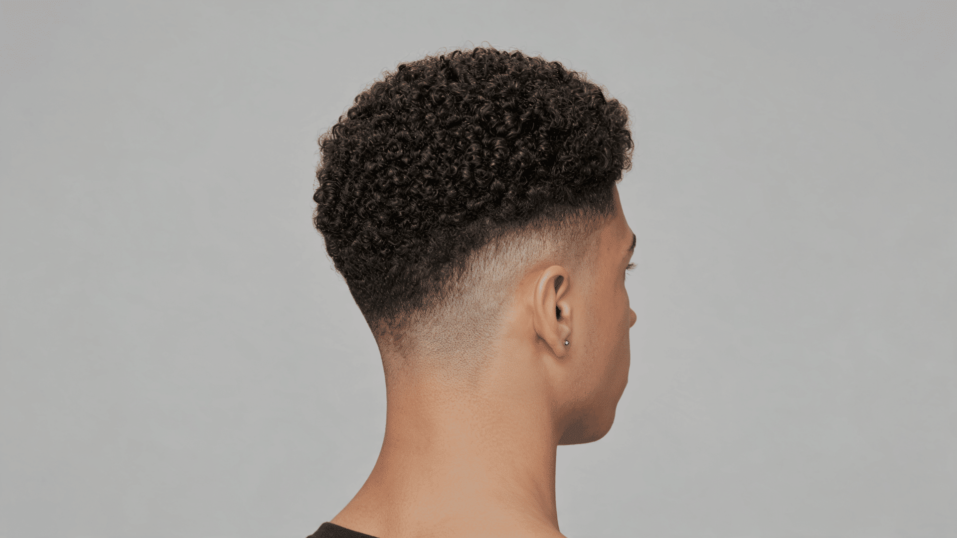 Side profile of man with curly top fade haircut, stud earring and blurred plain background