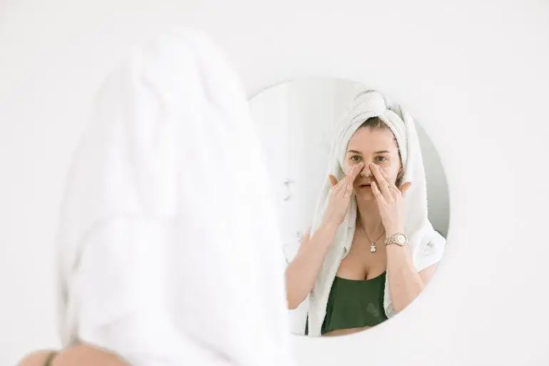 Woman applying skincare product in bathroom mirror with towel wrapped around head