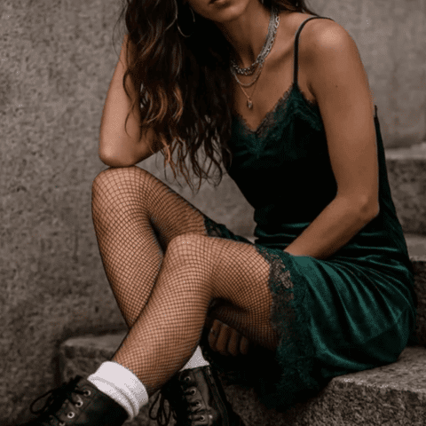 Tate McRae concert outfit idea: Woman sitting on stone steps wearing a dark green slip dress, fishnet tights, black boots, and layered necklaces.