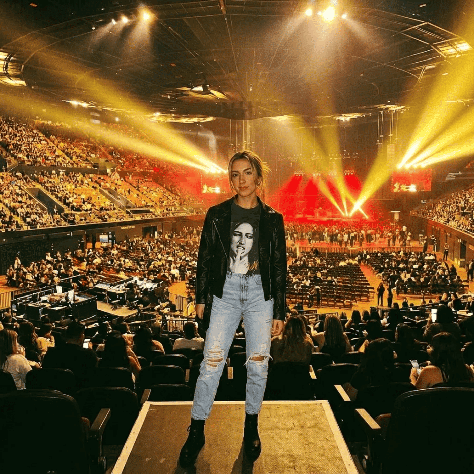 Tate McRae concert outfit idea: Woman standing in a concert arena with stage lights and audience behind her.