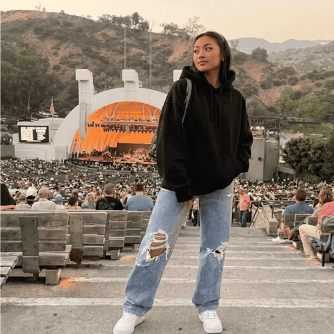 Tate McRae concert outfit idea: Woman wearing a black oversized hoodie, ripped light blue jeans, and white sneakers at an outdoor amphitheater concert.