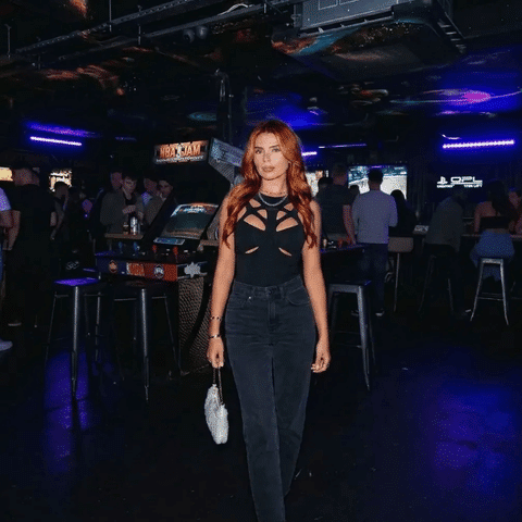 Tate McRae concert outfit idea: Woman wearing a black cut-out top, high-waisted black jeans, and holding a small white handbag in a dimly lit club or arcade setting.