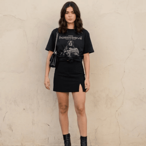 Tate McRae concert outfit idea: Woman wearing a black graphic t-shirt, black mini skirt with a side slit, black ankle boots, and a black shoulder bag standing against a plain wall.