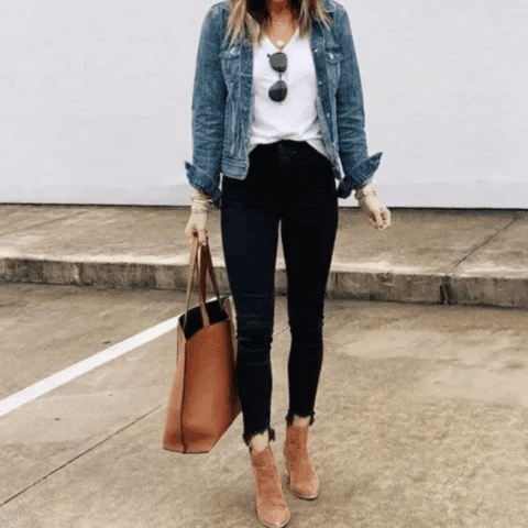 Tate McRae concert outfit idea: Woman wearing a white top, denim jacket, black skinny jeans, tan ankle boots, and carrying a brown tote bag while standing in a parking lot.
